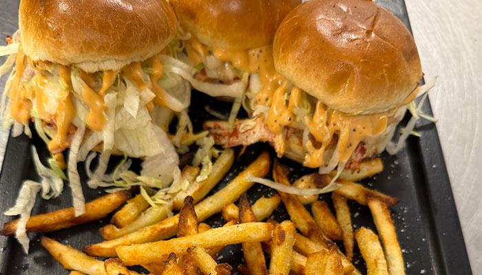Three sliders with lettuce and sauce, served with seasoned French fries on a black tray.